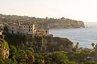 Tropea, Italy - 27.07.2023: Picturesque view of historic building perched on cliff, overlooking ocean and surrounded by lush greenery with distant cliffs and coastline in background [IBR123646525]
