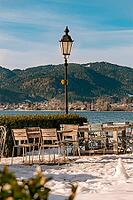 Outdoor area of a cafe on a snowy shore with a view of a lake and surrounding mountains under a blue sky, Tegernsee, Germany [IBR123895571]