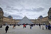 Paris, France - December 10, 2025: The ornate Renaissance facade and the iconic glass Pyramid of the Louvre Museum with multiple tourists scattered across the vast cobblestone courtyard [IBR123895594]