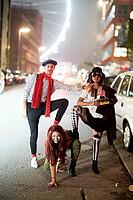 Dominance hierarchy of three friend in costume posing playfully under city lights on an urban street at night, nightlife, dog-like attitude of submissive playful woman crawling on street floor while her male friend steps on her back with his foot like her [IBR123999807]