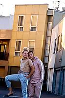 Couple posing playfully in a neighborhood street with colorful buildings, in front of apartments, real estate ownership, wearing casual clothes, sweater, coolness, cool, leisure, hanging ot, hip, trendy, fashionable, easygoing, lovers, lover, love, girlfr [IBR124192786]