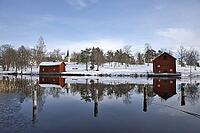 Classic red Swedish wooden house on the water with the reflection of the Borgvik church tower. Winter scene with snow and traditional Falunred architecture in Värmland, Sweden [IBR124200516]