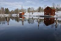 Classic red Swedish wooden house on the water with the reflection of the Borgvik church tower. Winter scene with snow and traditional Falunred architecture in Värmland, Sweden [IBR124200550]