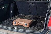 A single brown plastic case with silver clasps sits on a ribbed black rubber mat inside a car's cargo area, suggesting a journey or storage solution [IBR124491535]
