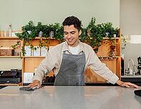 Smiling barista cleaning a counter in a cafe with a towel. Young male in an apron working as a bartender [IBR124491558]