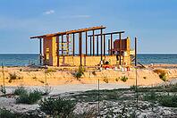 A brick building is under construction on the sandy beach near the sea, at sunset. Construction equipment can be seen [IBR124491563]