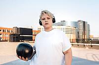 Confident female standing on the roof with medicine ball and headphones on her head [IBR124491575]