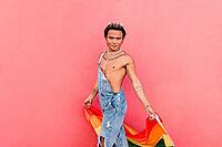 Young man walking with rainbow flag against pink wall [IBR124491598]