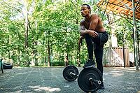 Bare-chest bodybuilder resting after deadlifting exercises at the sports ground. Young athlete stands with one leg on the barbell [IBR124491599]