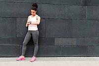 Young woman standing at wall, checking activity tracker [IBR124491625]