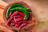 Bright green and red peppers are neatly placed in a round wooden bowl on a wooden board. The board has a natural finish, showing the grain of the wood [IBR124552189]