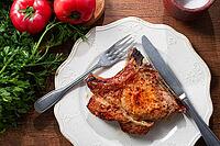 A pork chop rests on a white plate with a fork and knife. Fresh tomatoes and green herbs surround the plate on a wooden table [IBR124552205]