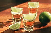 Three glasses with tequila and salt on the rims are arranged on a wooden table. Each glass has a slice of lime on top. A lime sits in the background under natural light [IBR124552248]