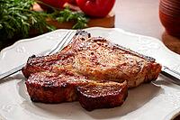 A pork chop rests on a white plate with a fork and knife. Fresh tomatoes and green herbs surround the plate on a wooden table [IBR124552264]