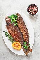 Two grilled fish are placed on a white dish. Fresh lemon slices and herbs are arranged beside them [IBR124585341]