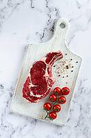 Raw rib eye steak, cowboy steak, on a white wooden chopping board, marble table, top view, no people [IBR124585364]