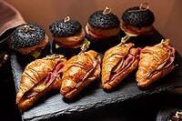 A selection of gourmet mini sandwiches featuring black sesame seed buns and croissants filled with deli meats and pickles. ideal for elegant catering events [IBR124626875]