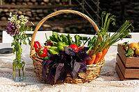 A wicker basket brimming with fresh vegetables like carrots, peppers, and greens sits on a table outdoors. nearby, a small vase holds wildflowers, creating a rustic and vibrant scene [IBR124626885]