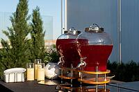 Two large glass dispensers filled with red juice are set up outdoors on a sunny day. the scene includes glasses and greenery, suggesting a refreshing outdoor gathering [IBR124626887]