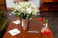 A wooden table elegantly set with a centerpiece of fresh daisies in a metallic vase. the table also features a single red rose, a lit candle, and neatly folded napkins, creating a cozy and inviting atmosphere [IBR124626899]