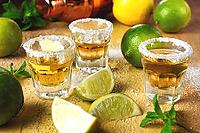 Several shot glasses sit on a wooden table with limes and mint leaves. A cocktail shaker is placed nearby. Slices of lime are scattered across the surface [IBR124629123]