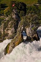 Rhine Falls in Neuhausen with rock formation in the middle of the waterfall, rock hill with hole above the water vortex, Swiss plume at the top of the rock formation with visitor platform, masses of water flow whirled up and foaming around the rock, green [IBR124636284]
