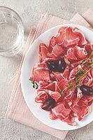 Freshly cut dried meat, served with olives and herbs, on a white plate, light background, no people [IBR124748955]