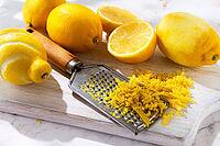 Lemons are placed on a wooden cutting board. Some lemons are whole while others are cut into slices. A zester is used to create lemon zest. The scene shows a bright kitchen [IBR124748966]