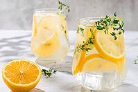 Two clear glasses hold a cold lemon drink with slices of lemon and sprigs of thyme. A half lemon rests nearby on a marble surface. Bright light illuminates the scene [IBR124748979]