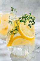 Two clear glasses hold a cold lemon drink with slices of lemon and sprigs of thyme. A half lemon rests nearby on a marble surface. Bright light illuminates the scene [IBR124748980]