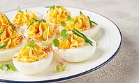 A plate holds several deviled eggs filled with yellow mixture and topped with spices and green garnishes. The dish is placed on a smooth light surface [IBR124748982]
