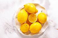 Lemons are arranged on a white decorative plate. One lemon is cut in half, revealing the bright yellow inside. The setting has natural light coming in [IBR124748986]