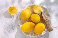 Four whole lemons sit on a white plate alongside a wooden juicer. The scene is bright with natural light, showing the lemons' color and texture clearly [IBR124748987]