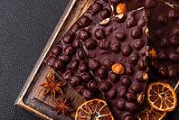 Food, Black bitter chocolate with whole hazelnuts, candied fruits and mint, broken into pieces on a dark concrete background [IBR124773737]