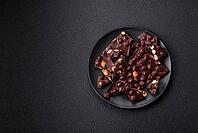 Food, Black bitter chocolate with whole hazelnuts, candied fruits and mint, broken into pieces on a dark concrete background [IBR124773748]