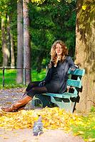 Young woman is sitting on the bench in autumn park and looking at camera [IBR124906366]