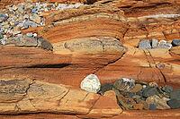 Detailed nature photo of a reddish-brown sandstone formation on a coast. The close-up shows the distinctive texture of horizontal sediment layers formed by centuries of erosion. Embedded in the rocky structure are various smaller, colorful pebbles and tid [IBR124920210]