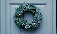 Simple holiday wreath made of pine branches and small white berries, hanging on a light gray wall. AI generated [IBR125000289]
