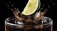 Extreme macro close-up of fresh lime wedge at precise millisecond of impact with dark cola rum cocktail surface inside clear highball glass with perfect crown splash forming thin translucent dark brown liquid columns around half-submerged lime trailing bu [IBR125000320]