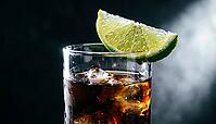 Extreme macro close-up of fresh lime wedge balanced diagonally on rim of tall highball glass filled with near-opaque dark cola rum cocktail, single Dedolight spot from right raking lime skin in full relief revealing every pore and oil cell, glass rim catc [IBR125000322]