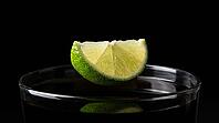 Extreme macro close-up of fresh lime wedge frozen mid-fall between two and four centimetres above perfectly still near-black rum cola cocktail surface inside tall highball glass, lime razor-sharp with every pore visible under raking Dedolight spot, undist [IBR125000324]