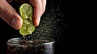 Extreme macro close-up of two fingers squeezing fresh lime wedge over tall highball glass containing dark rum cola cocktail, extraordinary subject is visible golden-green citrus oil micro-mist of ruptured oil cells hanging mid-air between lime and drink c [IBR125000325]