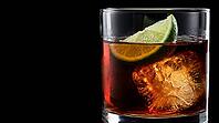 Macro close-up through clear glass wall of short rocks glass containing dark rum with lime wedge resting on large craft ice cube pressing slightly into its surface and dark rum filling to just above lime, Dedolight spot transmitting through glass turning  [IBR125000327]