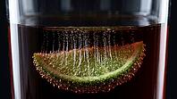 Macro view through clear glass wall of highball glass containing dark rum cola cocktail showing fully submerged lime wedge covered in dense cascading CO2 carbonation bubble trails erupting from every pore as nucleation sites simultaneously, Dedolight back [IBR125000328]