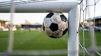 Football wedged in right-angle junction where white goal post meets white crossbar simultaneously touching both metal surfaces, viewed from inside goal looking outward with ball silhouetted against blurred green pitch and pale sky beyond, natural backligh [IBR125000329]