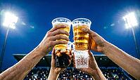Wide below-angle view looking up at four plastic cups raised simultaneously in diamond formation toast against open night stadium sky, held by four different wrist-level hands with two golden beer one dark cola with ice and one clear sparkling water, stad [IBR125000331]