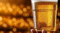 Extreme macro close-up of transparent plastic stadium cup filled with cold golden beer with dense condensation covering exterior surface showing individual droplets and rivulets in sharp detail, single hand visible at base, warm amber stadium bokeh fillin [IBR125000332]