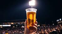 Single transparent plastic stadium cup of golden beer held at arm level against night stadium sky backlit by powerful overhead floodlight creating luminously translucent amber glow through liquid with condensation exterior dots as bright backlit points an [IBR125000333]