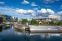 The modern Pole de Commerces et de Loisirs Confluence shopping and leisure center with its curved roof is reflected next to the green promenades of Quai Rambaud in the calm water of the Darse de la Confluence harbour basin under blue summer skies in Lyon, [IBR125002337]