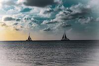 Two sailboats sailing on a tranquil ocean beneath a cloudy sky with a soft sunset glow. Aruba, The Netherlands [IBR125026133]